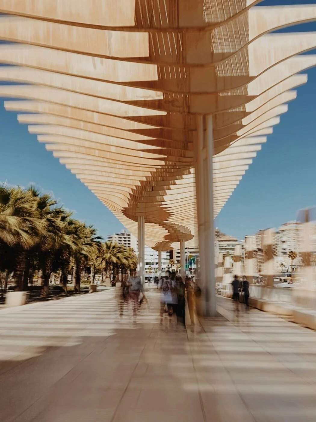 The wave-form wooden canopy along Malaga port, pedestrians blurred beneath it