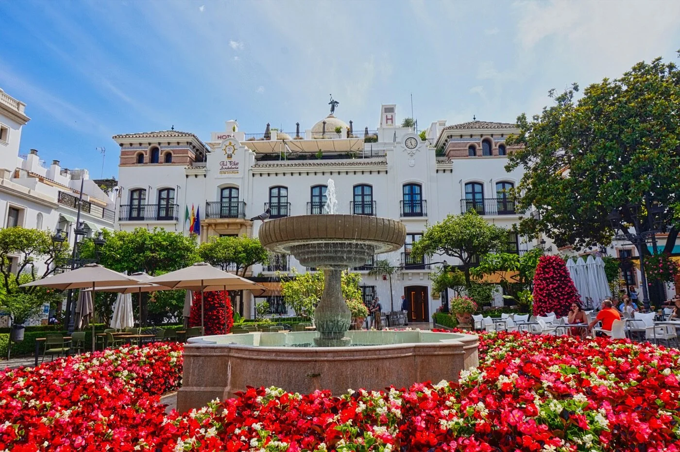 A sunlit plaza with a stone fountain, red flowerbeds and a whitewashed facade