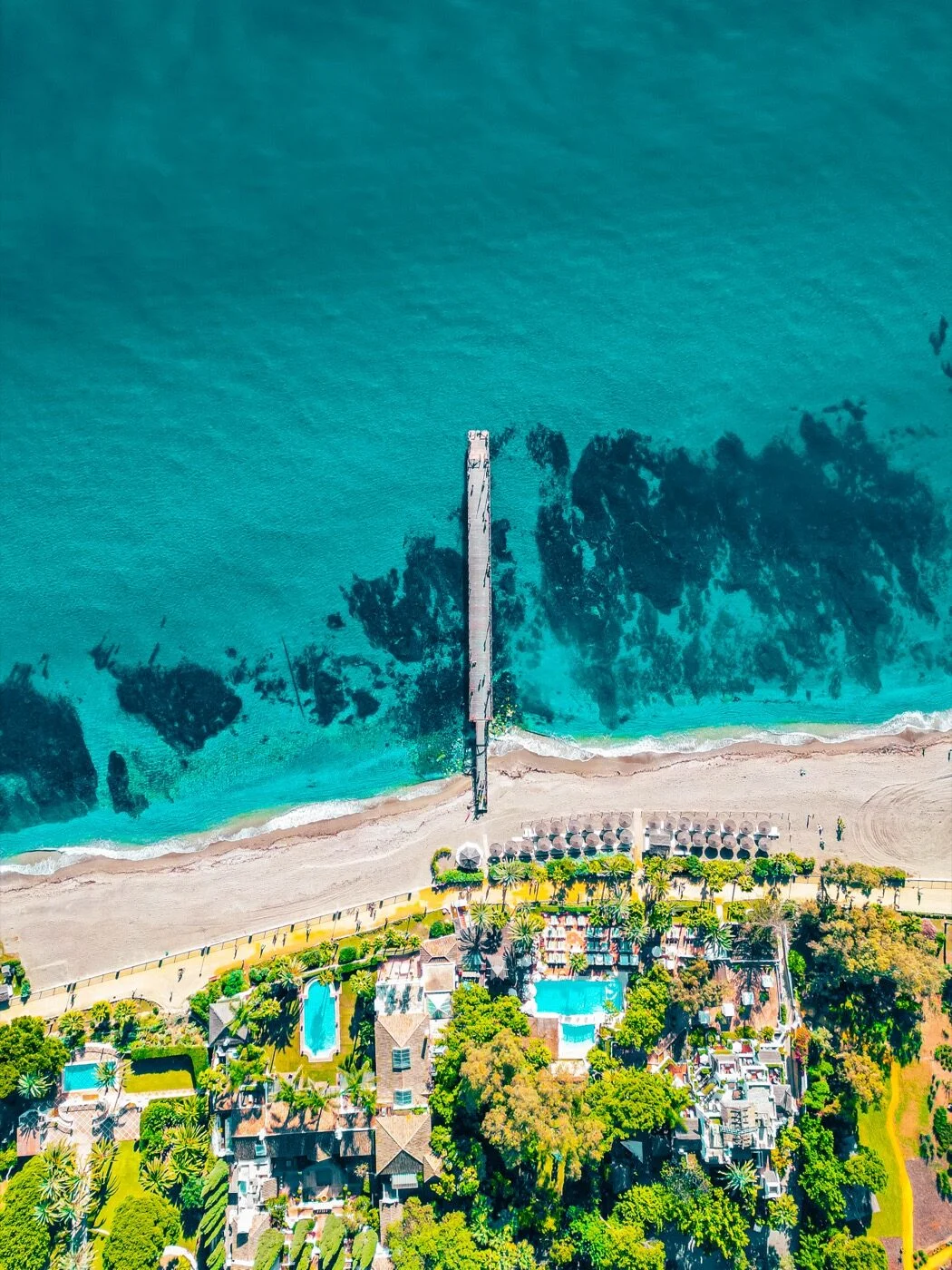 Aerial view of a wooden pier over turquoise sea, beach club pools and loungers below