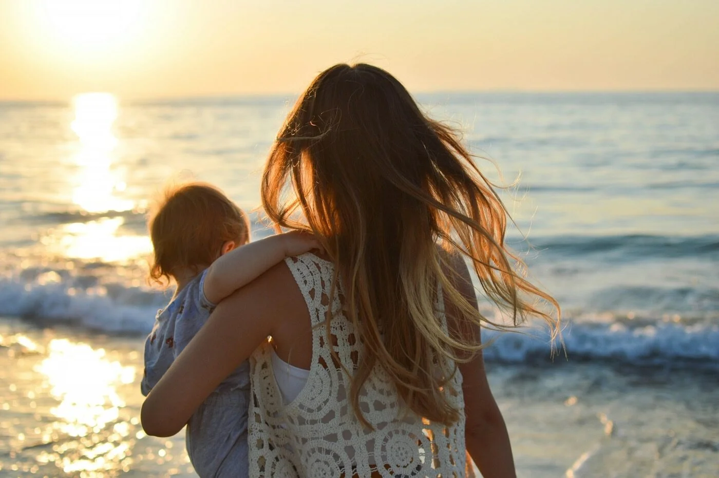 A mother carrying a toddler at the water's edge during a beach sunset